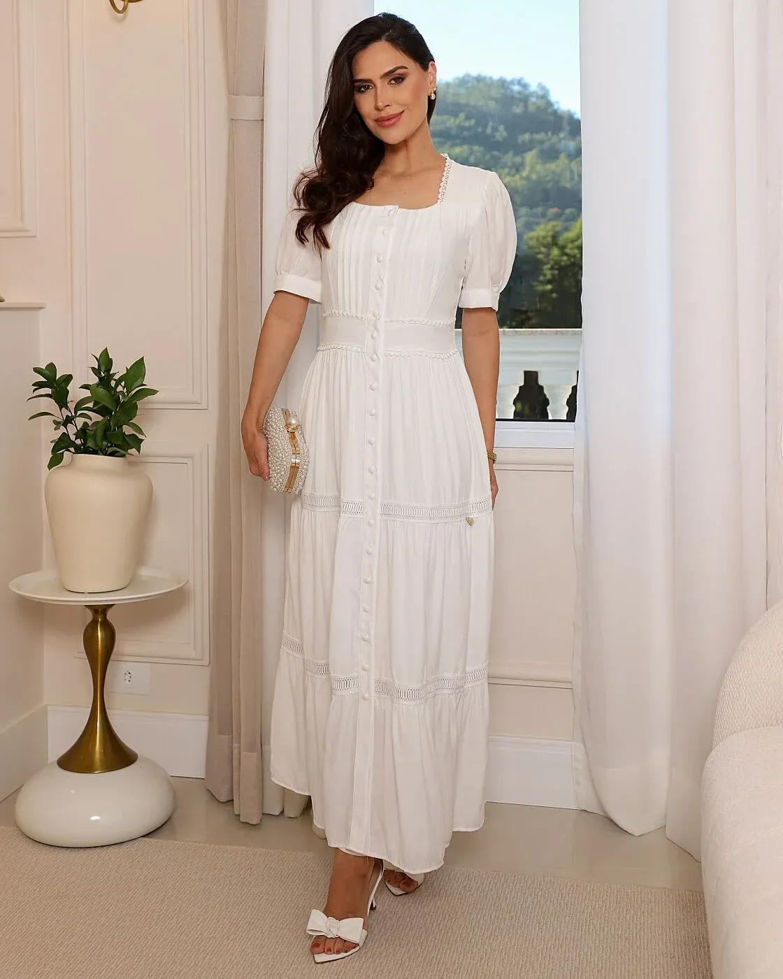 O Vestido Ágata, em um delicado tom branco, possui decote quadrado com acabamento em renda, mangas curtas levemente bufantes e pregas verticais no busto. A cintura marcada apresenta detalhes delicados, enquanto os botões encapados percorrem toda a frente. Sua saia longa e fluida possui recortes com renda entremeio e franzidos suaves, proporcionando movimento e leveza. Uma peça romântica, clássica e sofisticada, que transmite elegância e feminilidade.