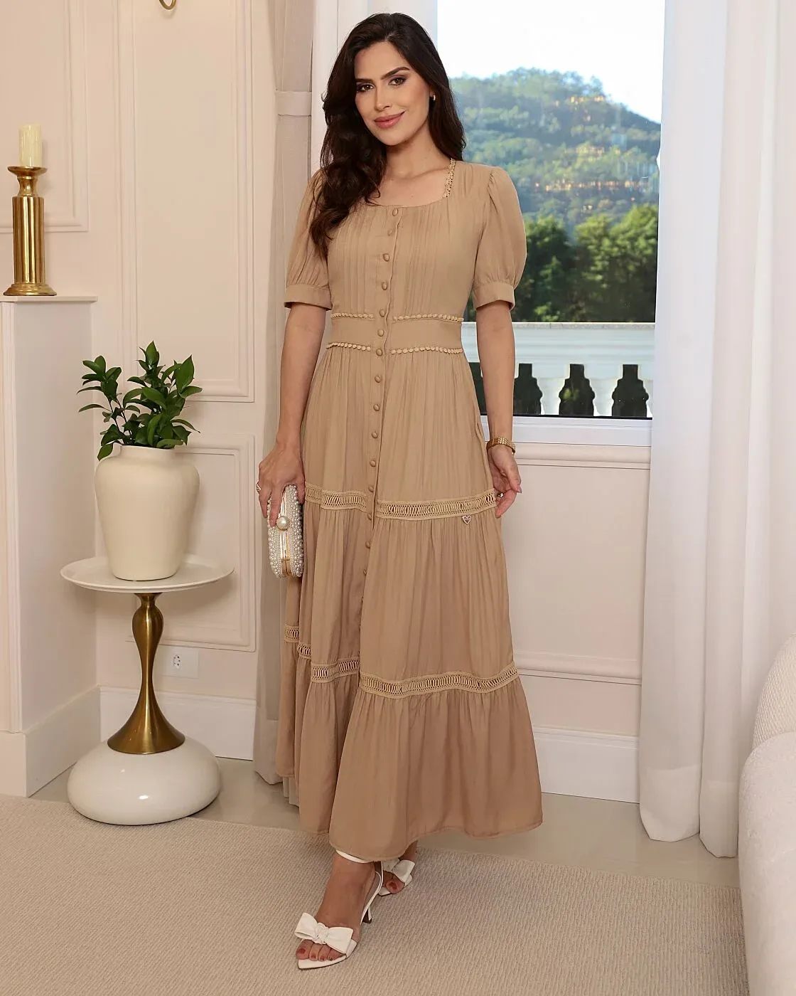 O Vestido Ágata, em um elegante tom bege, possui decote quadrado, mangas curtas levemente bufantes e delicadas pregas verticais no busto. A cintura marcada apresenta detalhes em acabamento rendado, enquanto os botões encapados percorrem toda a frente. Sua saia longa e fluida conta com recortes, franzidos e aplicações de renda entremeio, proporcionando movimento e delicadeza. Uma peça romântica, clássica e sofisticada, perfeita para quem ama elegância e feminilidade.